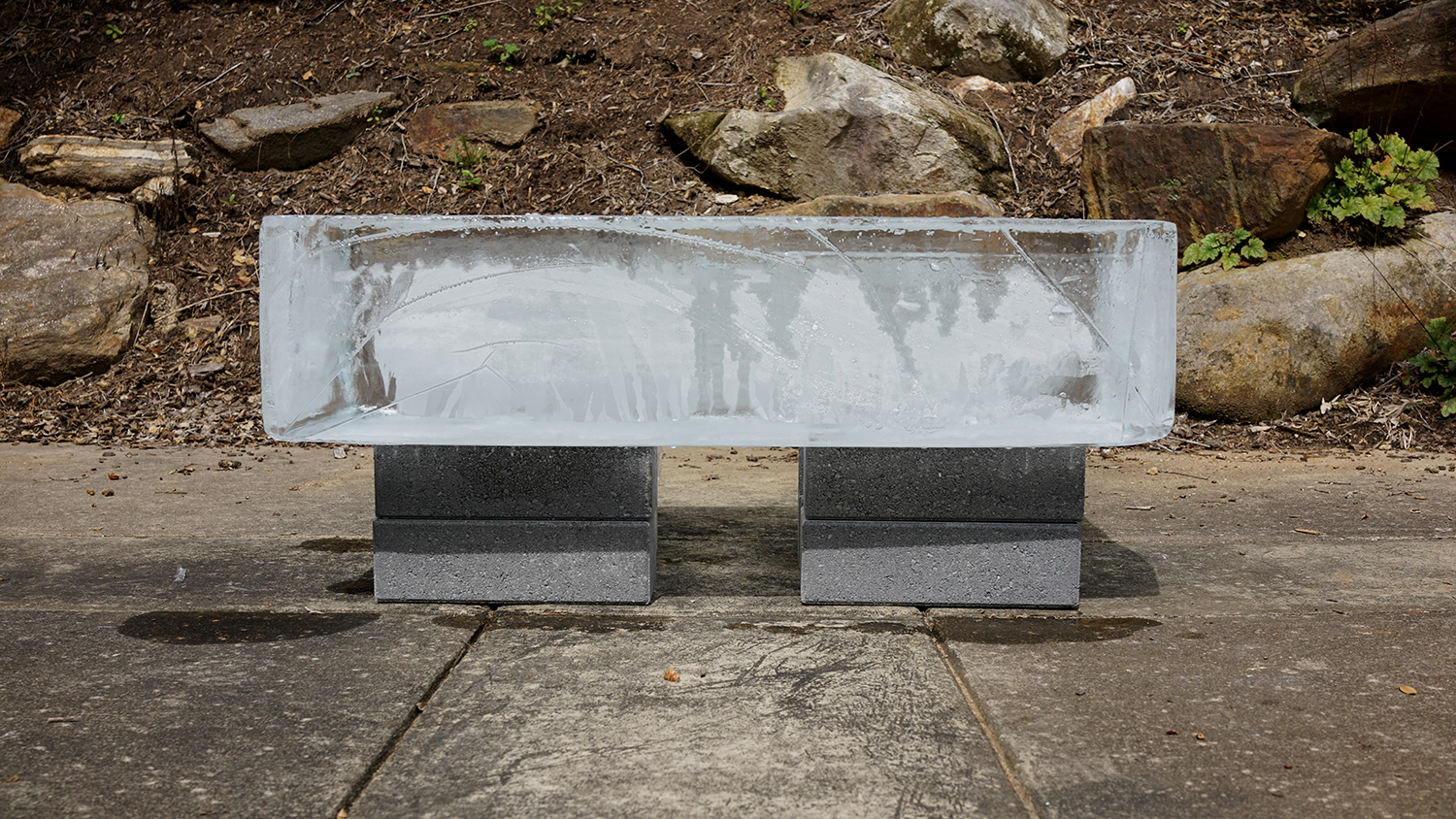 All That Is Solid Melts Into Air, 2025, 40 x 18 x 20 bench (300 lb ice block)<br><br>Describing how the bourgeoisie preserve control Karl Marx wrote “all that is solid melts into air.” New becomes old before it can solidify. People constantly experience changing realities and truths. Uncertainty cloaks the facts. Dressed in a suit and tie, similar to John Cage in his 1959 “Water Walk,” I sit on a bench of ice. ReViewing Black Mountain College Conference attendees partake too. Accompanying the performance, a speaker broadcasts the sound of melting ice. In “Water Walk,” Cage created music from sounds of overlooked everyday water related items, while “All That Is Solid Melts Into Air’s” soundscape makes audible the almost imperceptible melody of melting ice. Wet bottoms from the melting ice imprint and remind us of our experience of unrelenting change and ambiguity.