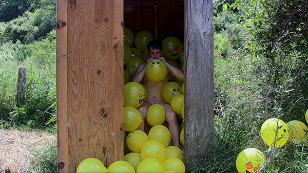 Take My Breath Away, 2018, video still<br><br>Go to menu, click VIDEOS, to watch excerpts<br><br>I perform with Emoji balloons to examine the reduction of language and communication to mere symbols. Entering a privy, I sit and inflate, using my breath, 100 yellow Emoji balloons. My intent was for the balloons to fill up the space around me. But like life, intention yields to reality. Unleashing each balloon, it casually floats to a new resting spot. Associations proliferate between the elements. The comical sputter of the balloons reverberates noises made by the body during waste elimination.